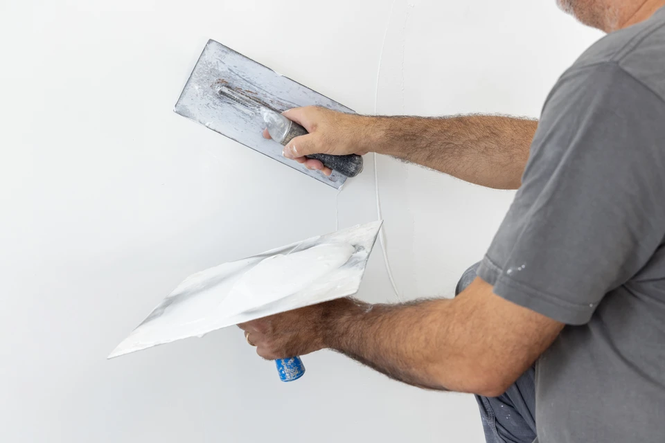 Int&eacute;rieur d'un chantier de r&eacute;novation avec ouvrier appliquant un enduit ou peinture blanche au pinceau sur une surface murale